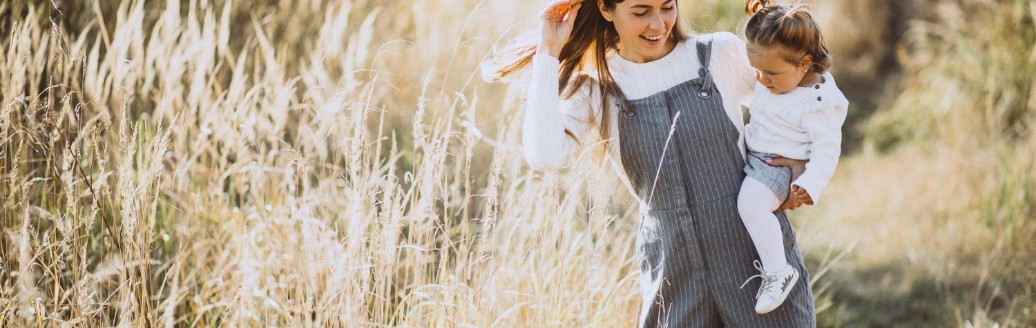 Eine junge Frau in Latzhose hat ein kleines Mädchen auf dem Arm und spaziert über ein Feld mit hohem Gras.
